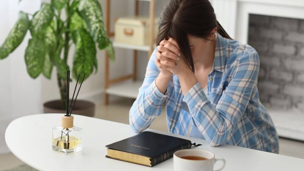 woman sitting at a desk with hands folded as she prays over her bible