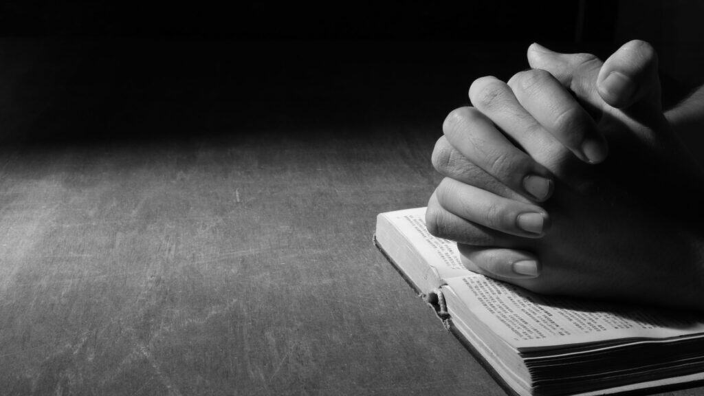 black and white image of clasped hands laying on top of an open Bible